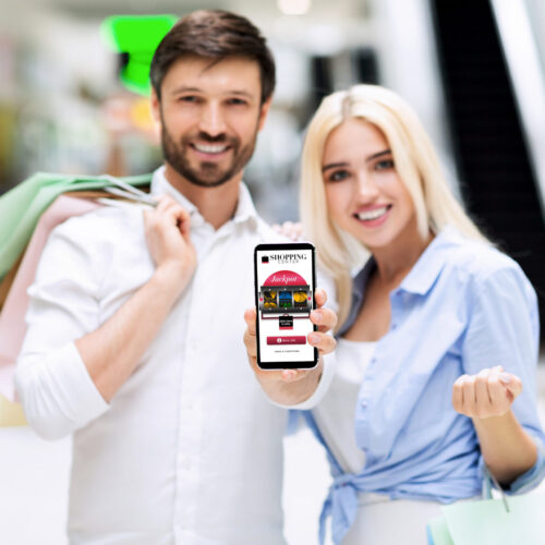 Shopping App. Smiling Couple Showing Blank Cellphone Screen Standing In Mall Center. Mockup, Shallow Depth